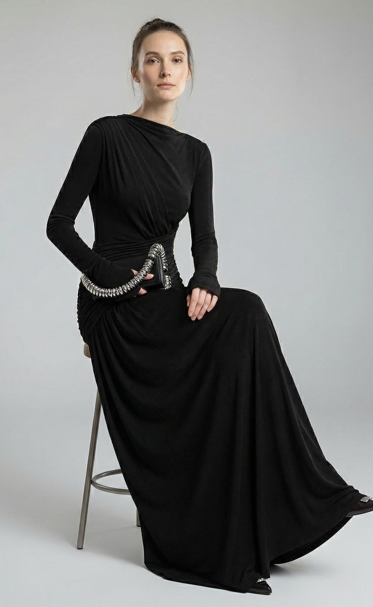 Black high-neck maxi gown worn seated on a stool, the model holding a small bag with a chunky chain handle.
