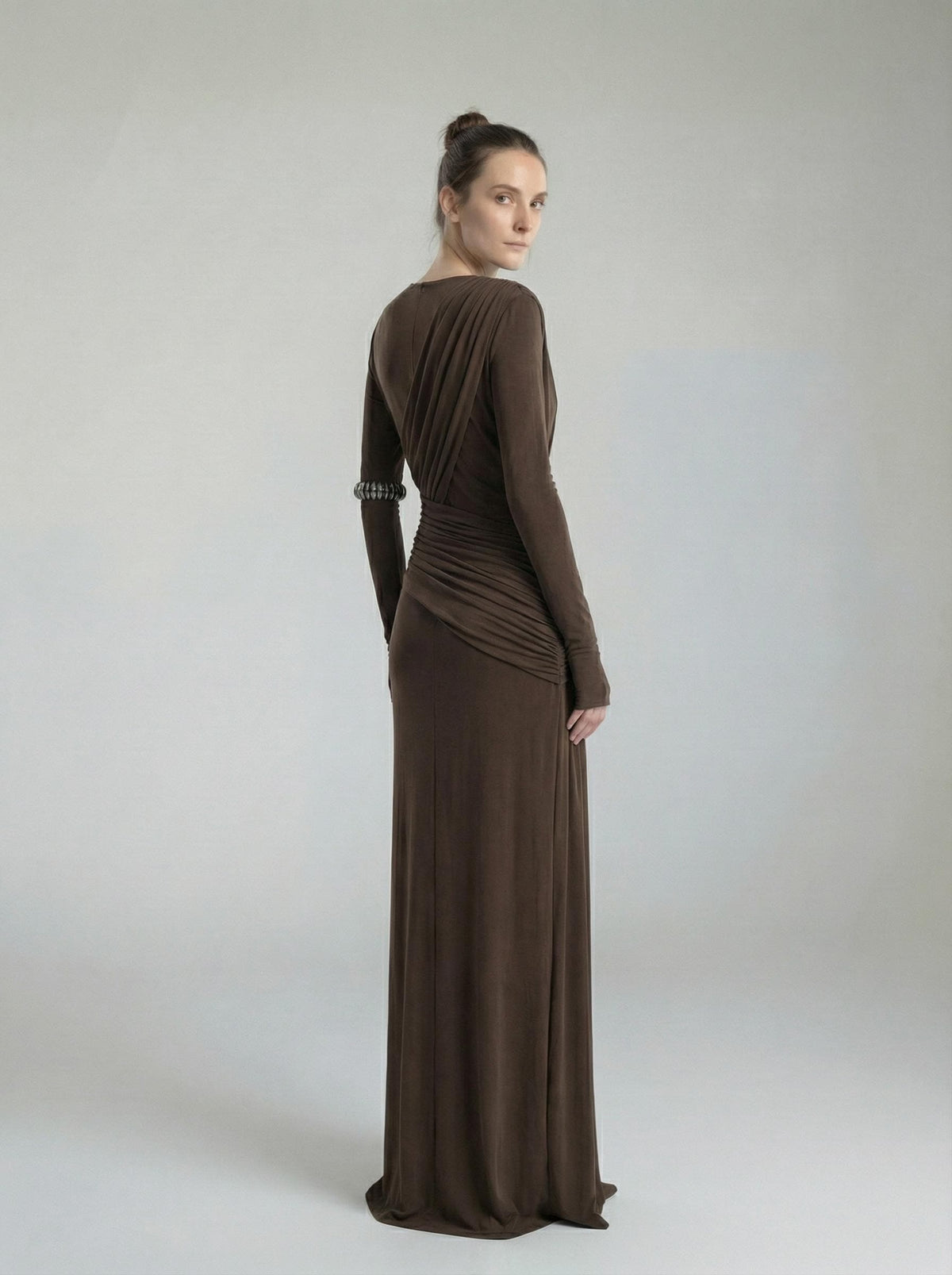 Brown high-neck maxi gown viewed from the back, the model looking over her shoulder in a plain studio.