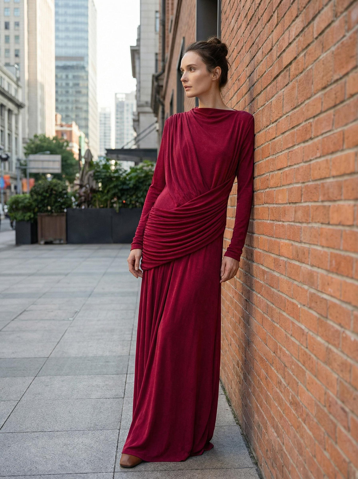 Red high-neck maxi gown with draped bodice and ruched waist, worn leaning against a brick wall on a city street.