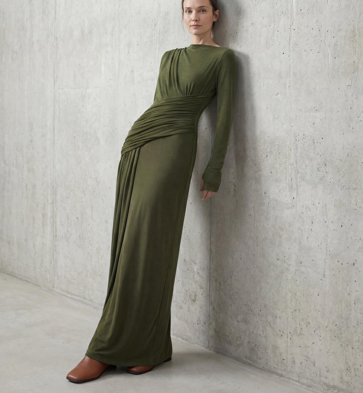 Olive Green high-neck maxi gown worn leaning against a bare concrete wall, shown full length from a three-quarter angle.