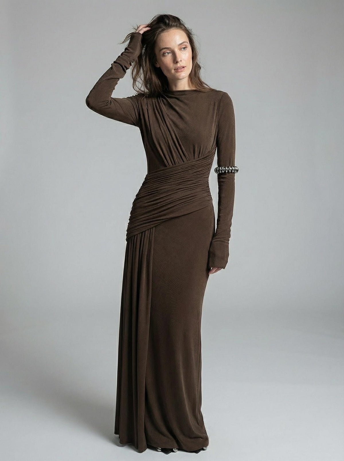 Brown high-neck maxi gown seen from the front at full length, the model raising one hand to her hair.