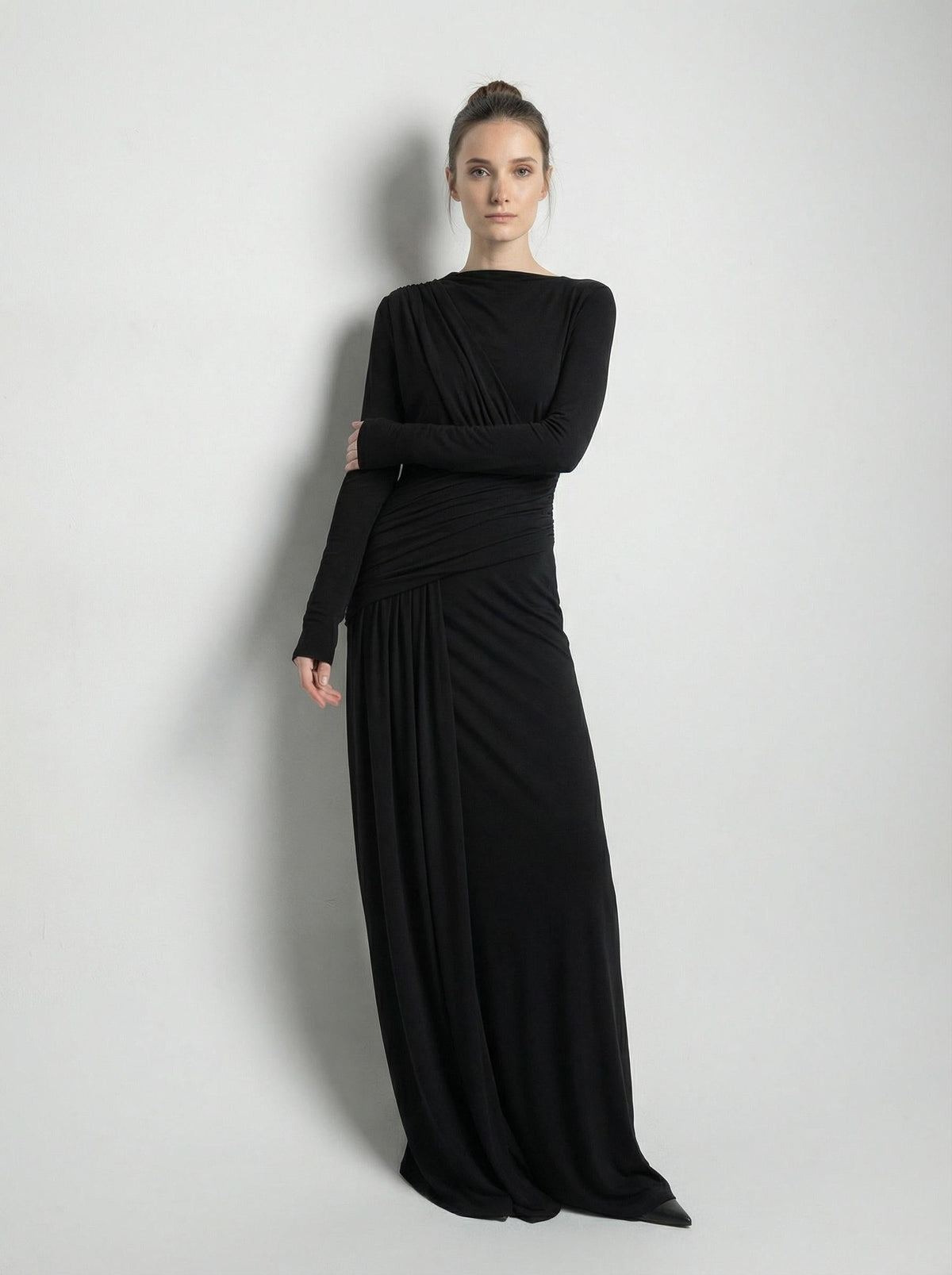 Black high-neck maxi gown shown front on at full length with arms folded, against a plain pale wall.