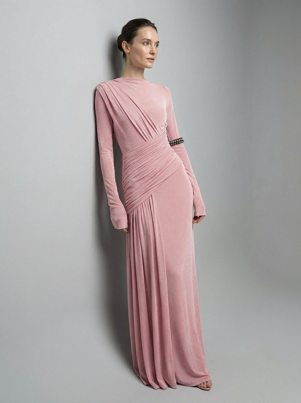 Pink high-neck maxi gown from a three-quarter angle against a plain wall, showing the draped shoulder and ruched waist.